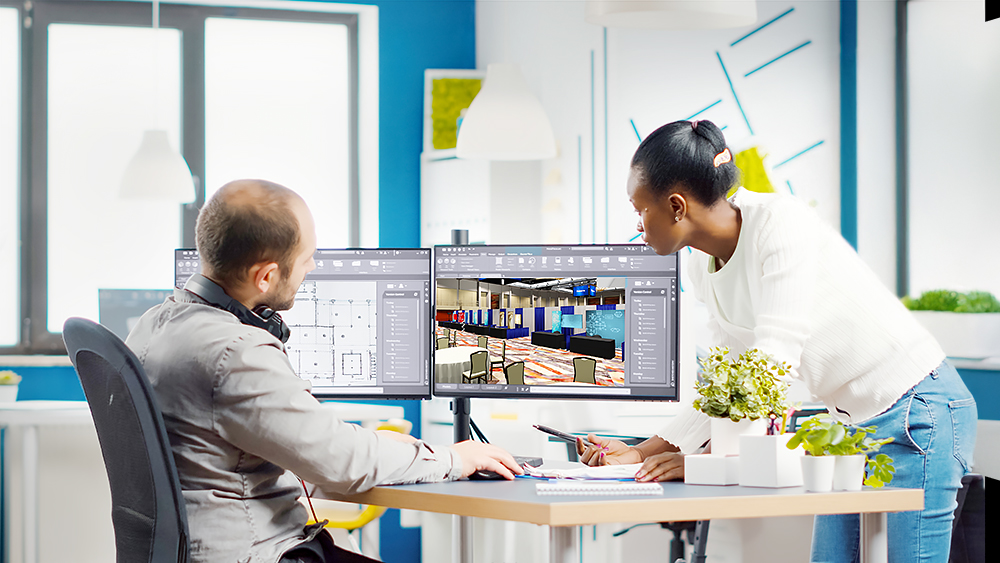 A man sitting at a desk and a woman standing beside him collaborate on interior design plans displayed on dual computer monitors in a modern office.