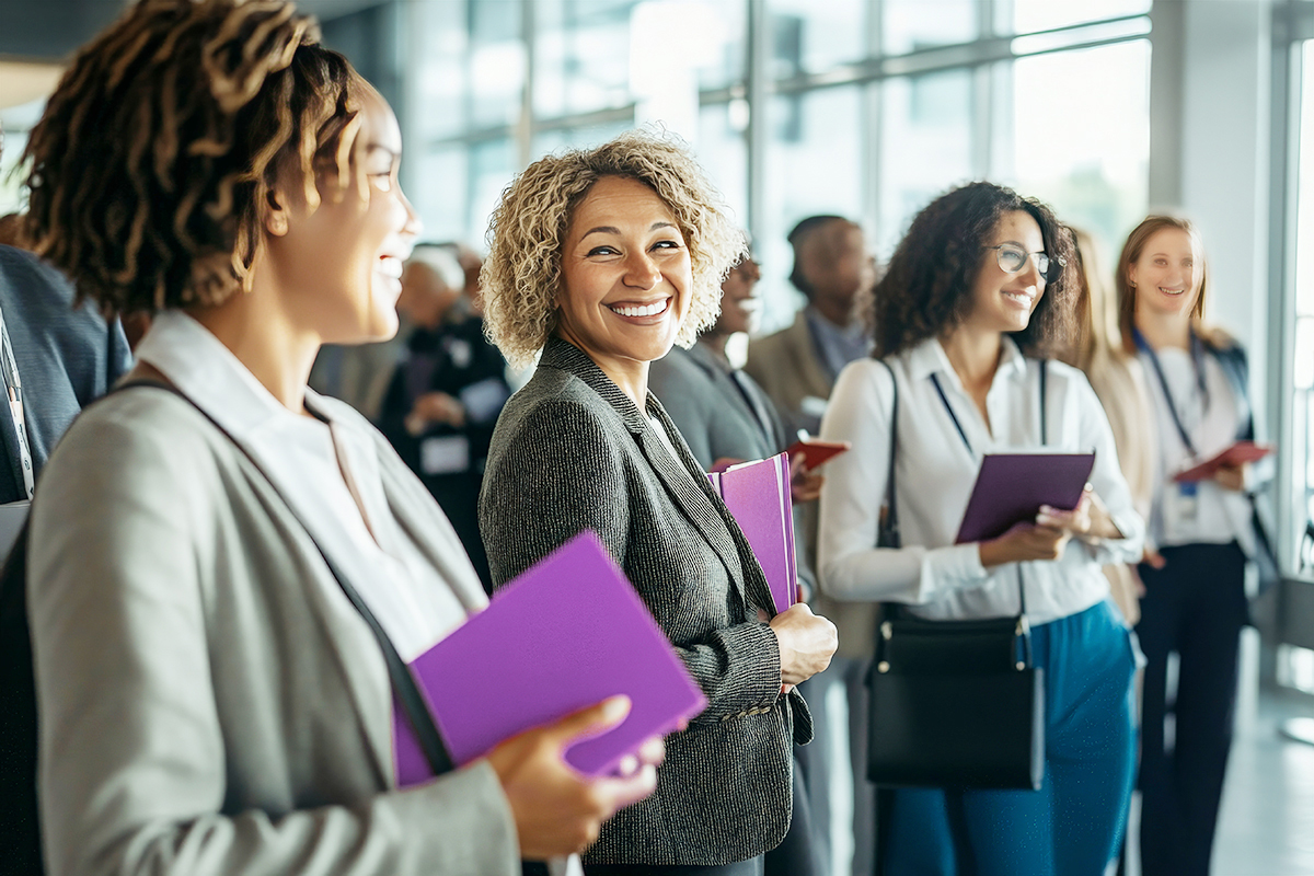 A group of professionally dressed people stand together in a bright, modern office. Several women in the foreground smile and hold purple folders, appearing engaged and happy.