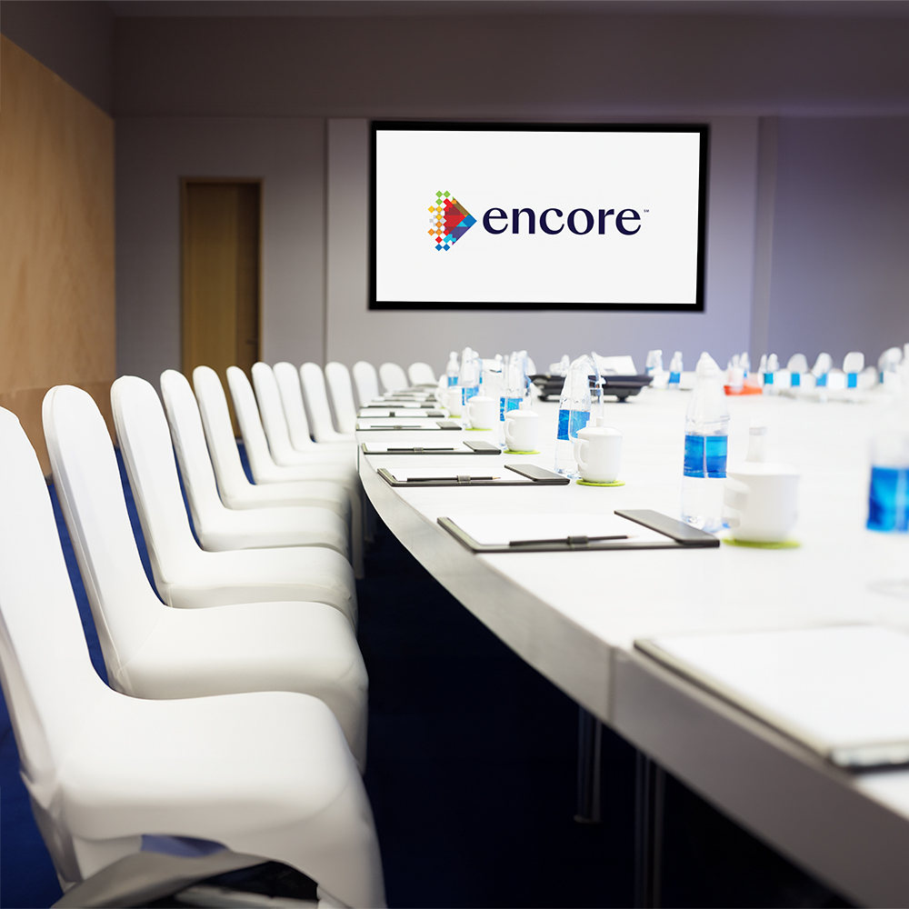 A modern, empty conference room with a long table set with notepads, pens, water bottles, and cups. White chairs line both sides, and a screen at the front displays the “Encore” logo.