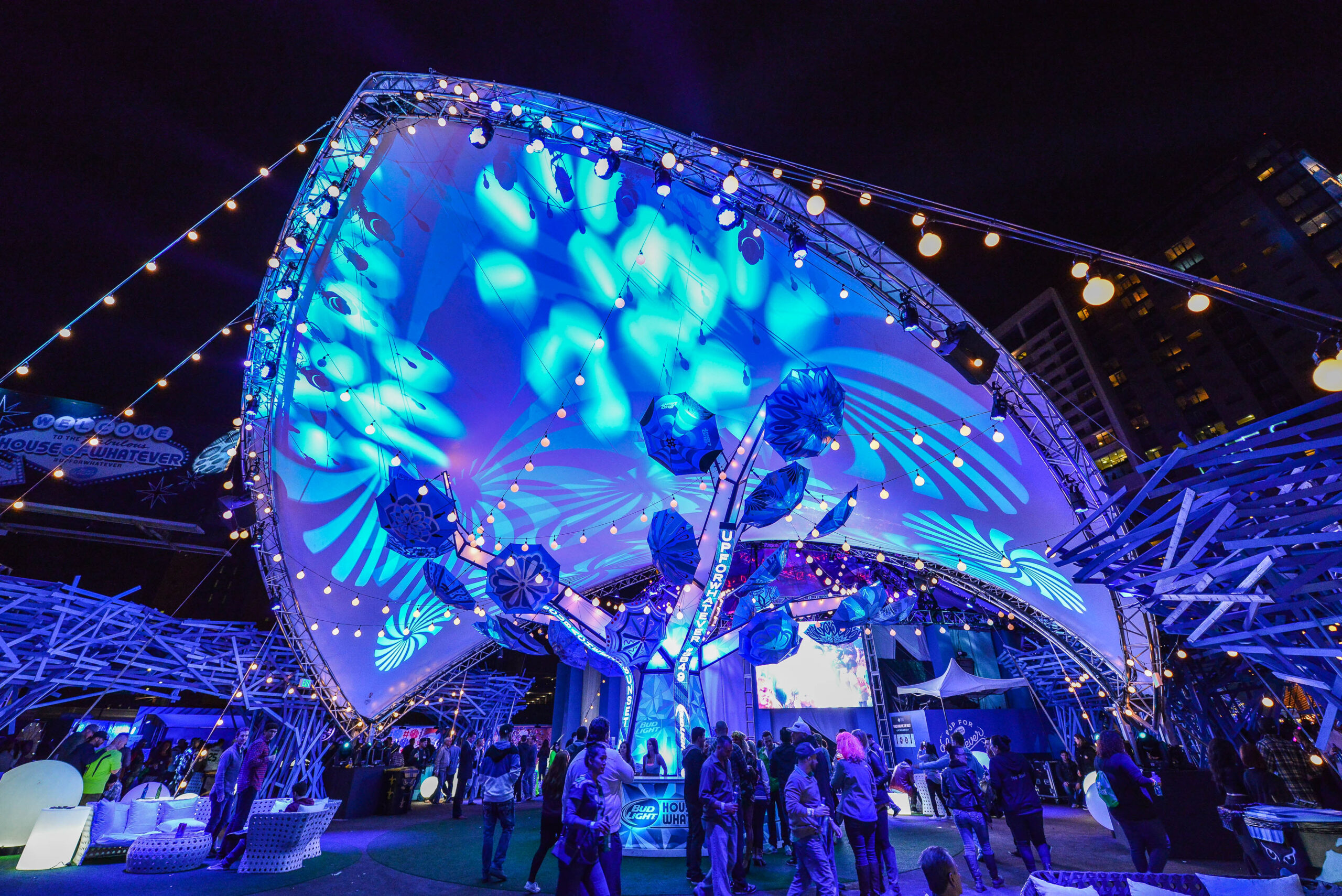 A crowd gathers under a large, futuristic canopy illuminated with blue lights and intricate patterns at night. The scene is festive, with people mingling and urban buildings visible in the background.