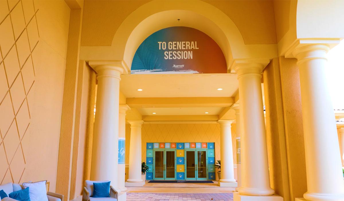 Wide archway with columns at a Marriott hotel; a sign above reads “TO GENERAL SESSION.” Behind the arch, glass doors and colorful square decals are visible; blue and white cushions sit on the left.
