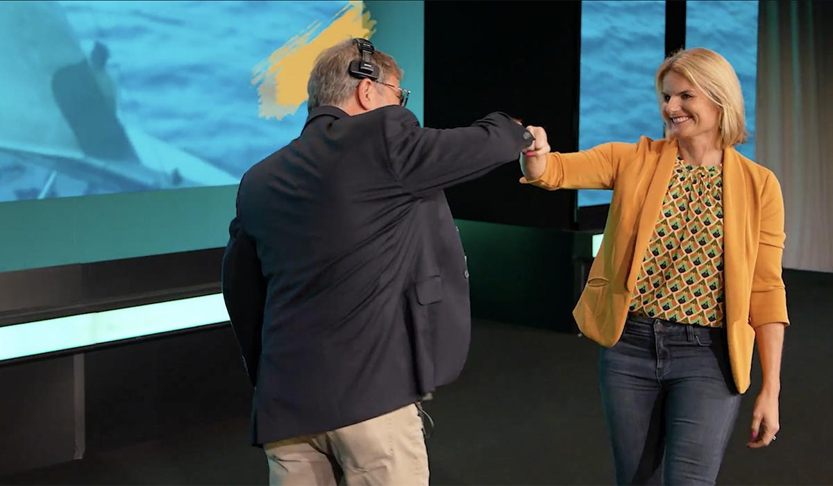 A man and a woman smile and greet each other with a fist bump on a stage. The woman wears a yellow blazer and patterned shirt; the man wears a dark blazer and headset. A blue-toned screen is visible in the background.