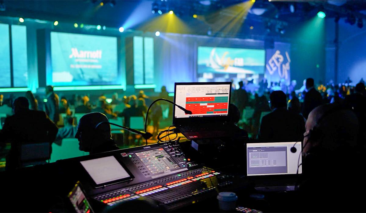 Audio-visual technicians operate control panels and computers at the back of a dimly lit event venue, with colorful stage lights, large screens, and an audience in the background.