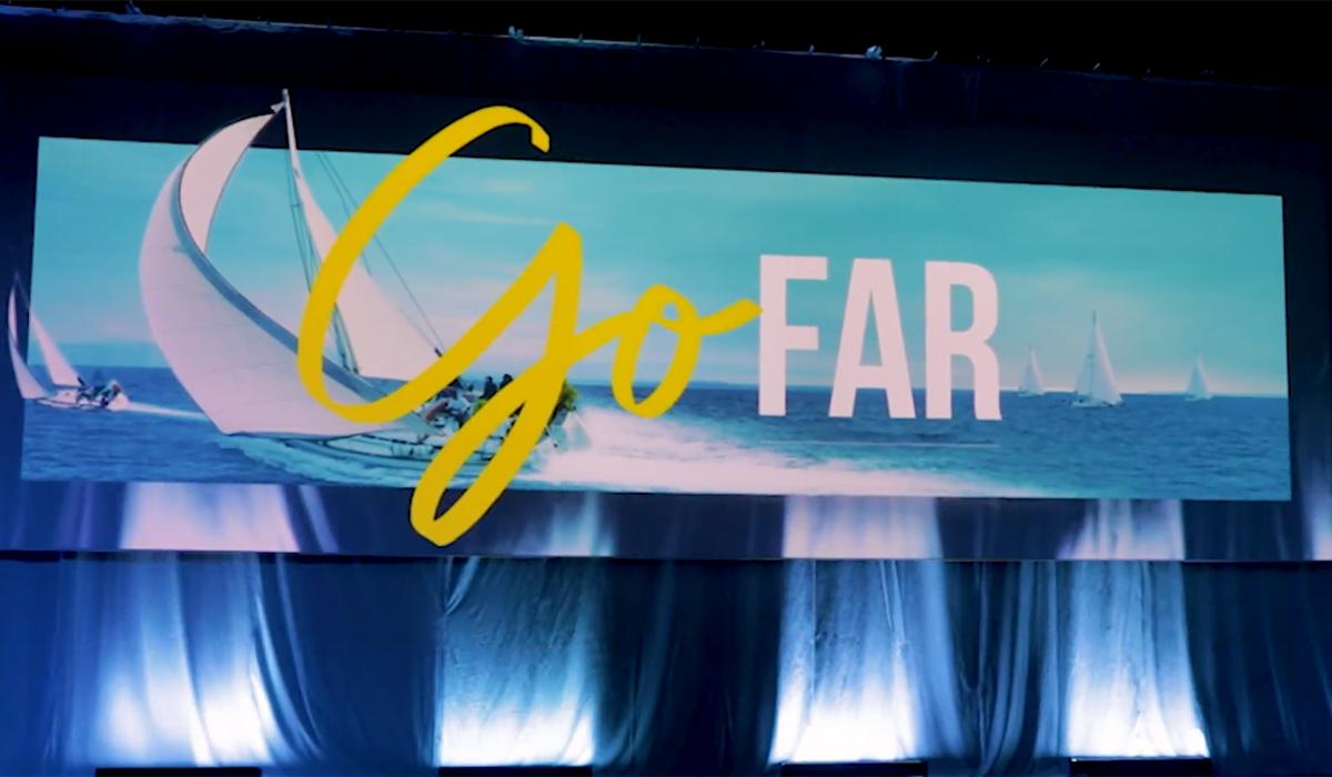 A large banner with a photo of sailboats on the ocean and the words Go FAR in bold, with Go in yellow script and FAR in white capital letters. The banner is displayed above a stage with blue lighting.
