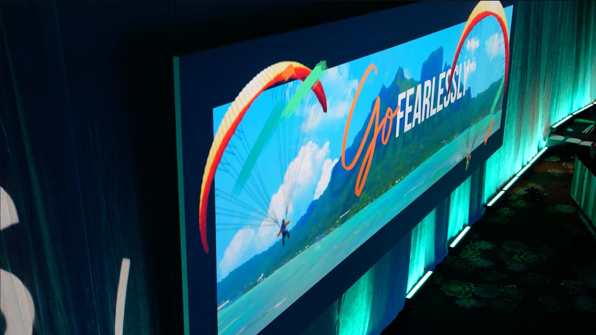 A large screen displays an image of a paraglider over mountains and water, with the words Go FEARLESSLY written in bold, cursive text across a blue sky with clouds.