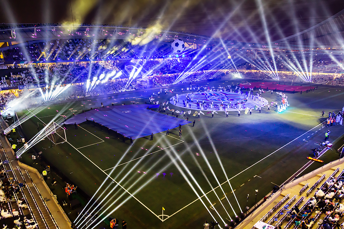A brightly lit stadium filled with spectators hosts an elaborate event with spotlights, performers, and colorful displays on the field, creating a festive atmosphere.