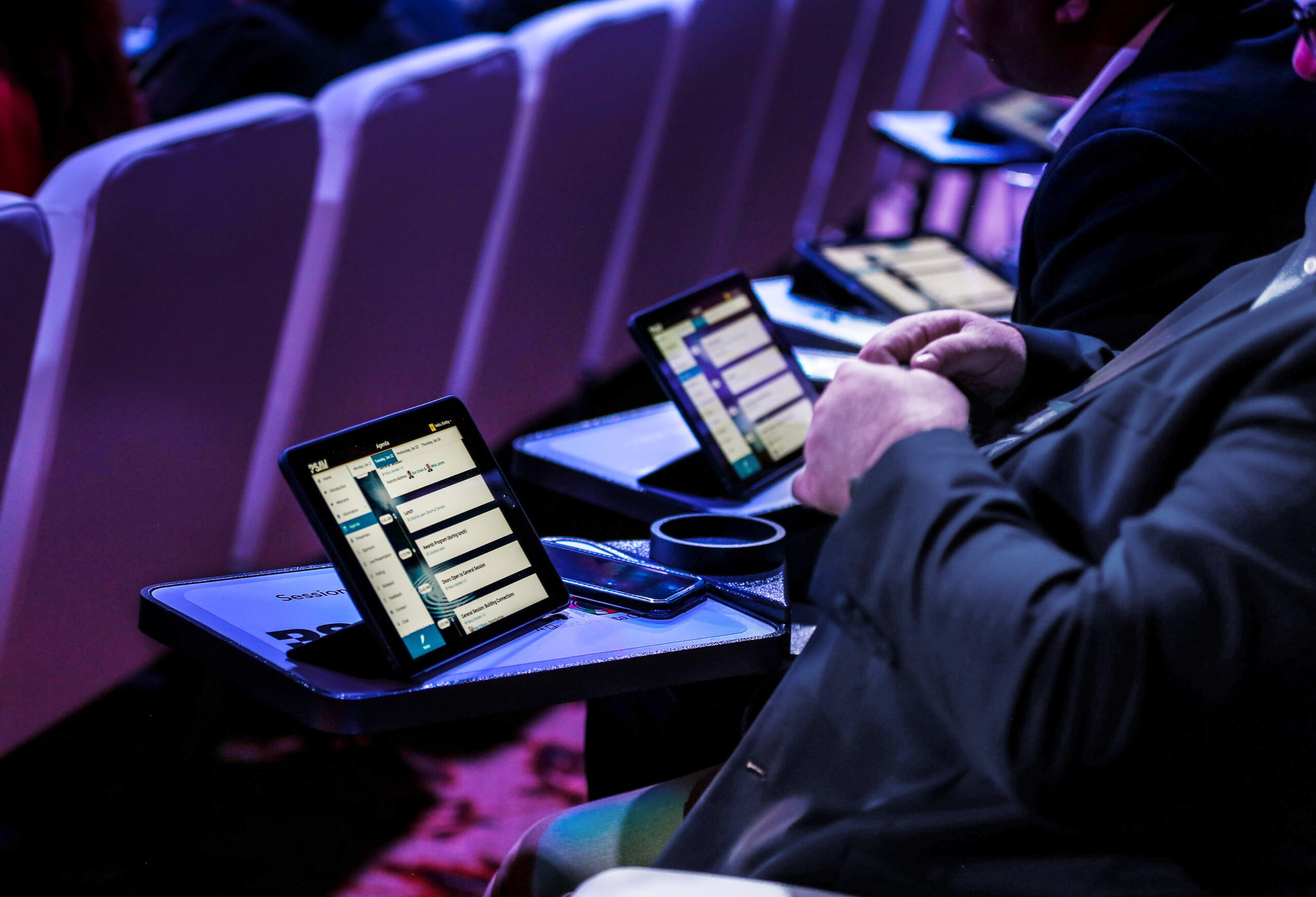 People seated in a row at an event, each with a tablet device on a small tray table, displaying apps and documents, in a dimly lit, high-tech environment.