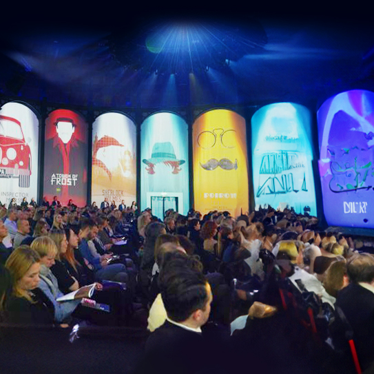 A large audience sits in a darkened theater, facing a brightly lit stage with tall vertical screens displaying colorful, stylized graphics and text. The atmosphere appears lively and engaging.