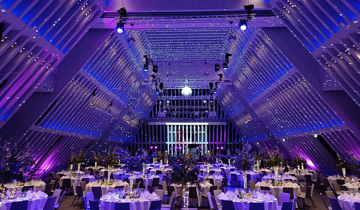 A large, modern banquet hall with tall, angled walls, decorated with purple and blue lights, twinkling string lights overhead, and elegantly set round tables for a formal event or celebration.