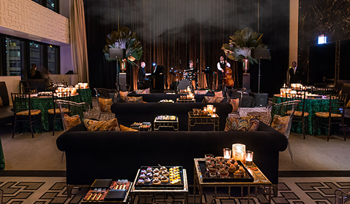 Elegant lounge set up for an event with black sofas, candlelit tables of desserts, and a jazz band performing on stage in the background; large plants decorate the stage area.