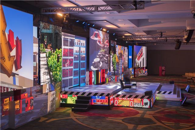 A large stage with colorful, vertical panels displaying various images and patterns, including street signs and cityscapes, is set up in a spacious event hall with rows of empty chairs in the background.