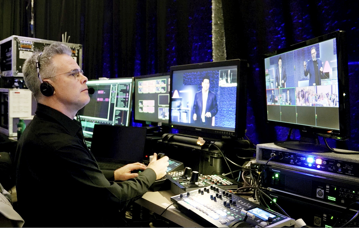 A man wearing headphones operates audio-visual equipment at a control station, with multiple screens displaying a person speaking on stage and various event footage in a backstage or broadcast environment.