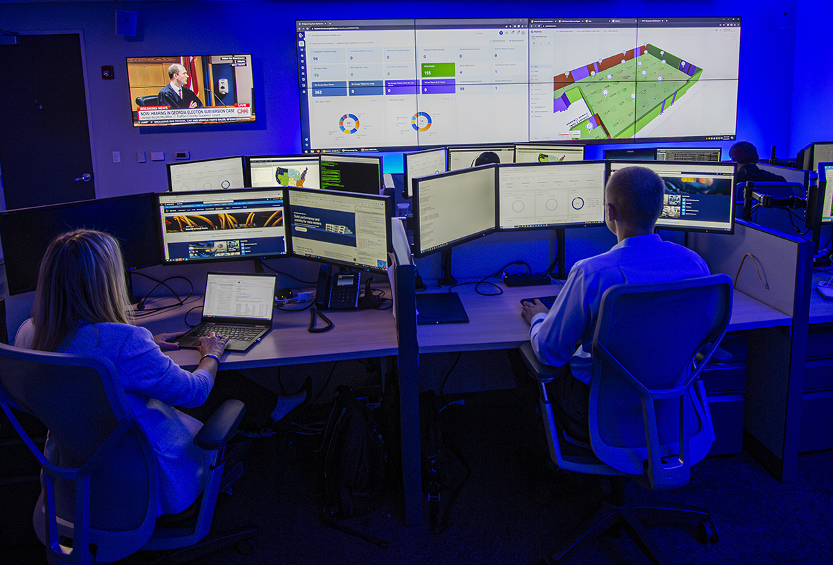 Two people sit at a control center desk with multiple computer monitors displaying data, charts, and a news broadcast. Large screens on the wall show digital dashboards and a virtual environment, all under blue lighting.