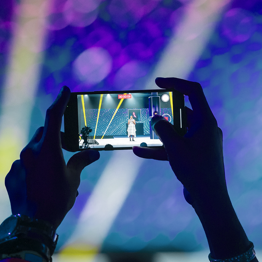 A person holds a smartphone, capturing a photo or video of a performer on a brightly lit stage with colorful, blurred lights in the background.