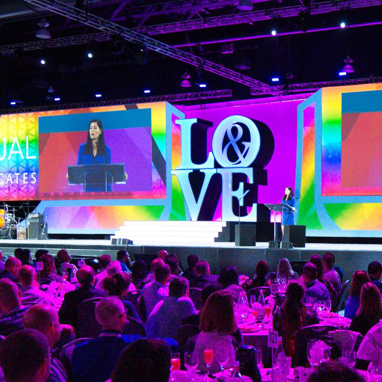 A woman speaks at a podium on a brightly lit stage with a large LOVE sign behind her. Audience members are seated at round tables in a darkened room, watching the presentation.