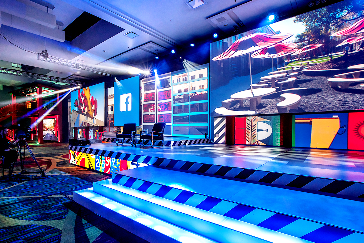 A vibrant, colorful stage setup with bright lights, digital screens displaying Facebook and outdoor scenes, and several empty chairs arranged for a presentation or event.