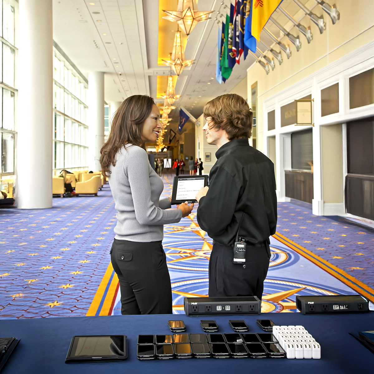 Two people stand talking in a brightly lit hallway with flags and star-shaped lights overhead. In the foreground, a table displays various electronic devices, including phones and tablets.