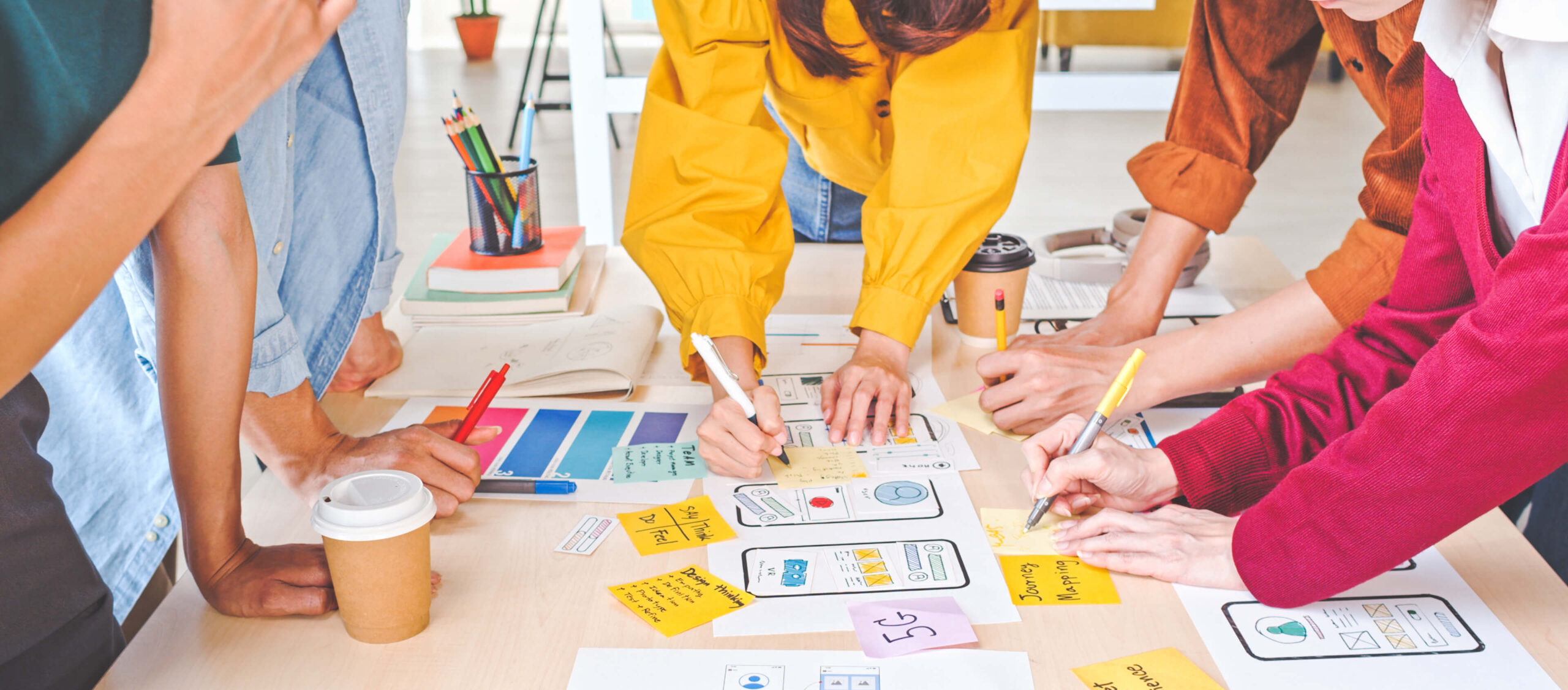 A group of people collaborates at a table covered with papers, wireframes, sticky notes, and coffee cups, brainstorming and sketching ideas for a mobile app design.