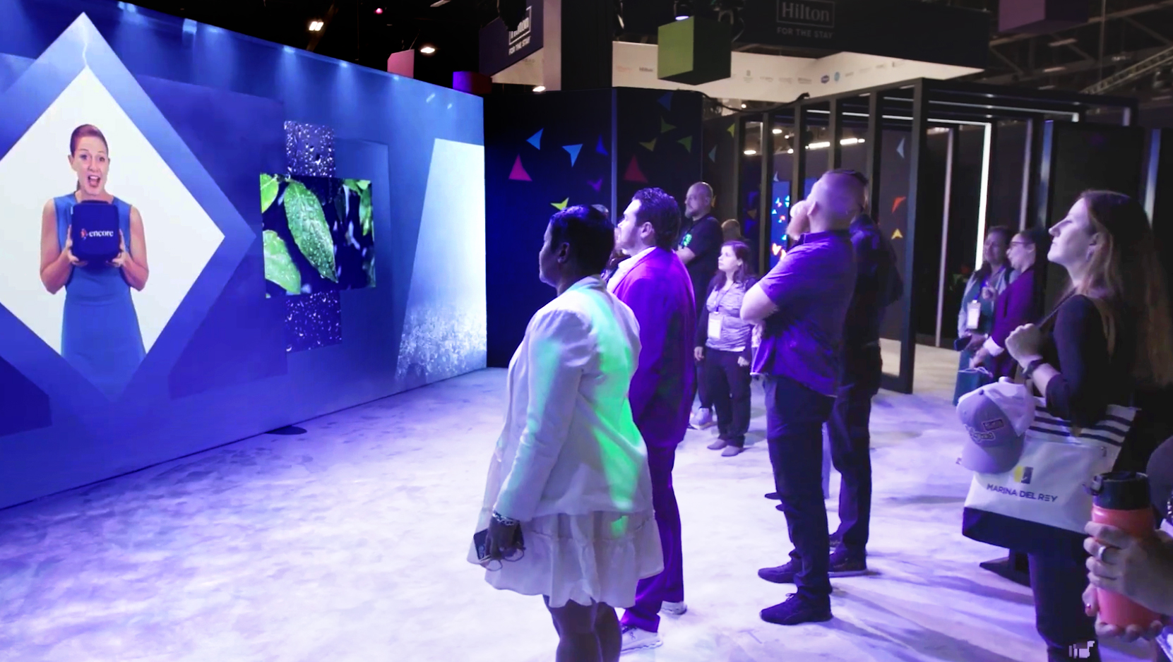 A group of people stands in an exhibit space, watching a large interactive digital screen featuring a woman speaking and animated graphics. The scene is brightly lit with colorful displays.